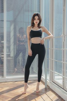 Woman in a black sports bra and leggings standing barefoot by a window