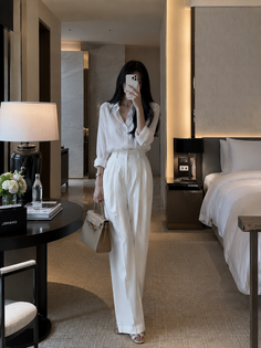 Woman in a white shirt and wide leg pants taking a mirror selfie in a hotel room