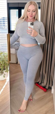 Woman with long blonde hair in a grey crop top and leggings taking a mirror selfie