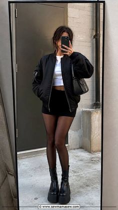 Woman in a black bomber jacket and mini skirt taking a mirror selfie outdoors