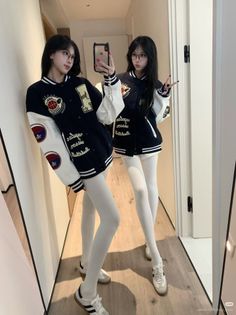 Two women in varsity jackets and white tights taking a mirror selfie indoors