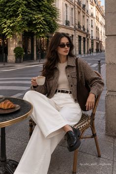 Woman in a plaid jacket and white pants sitting at an outdoor cafe with a croissant