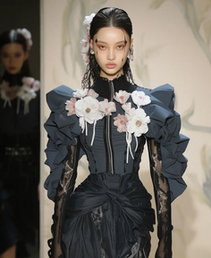 Runway model in a dark dress with floral embellishments and dramatic sleeves