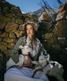Woman with long curly hair holding two baby goats outdoors by a stone wall