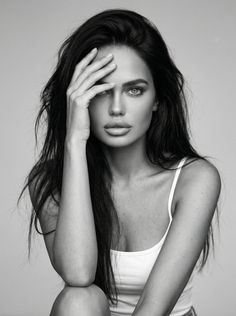Woman with long dark hair in a white tank top, hand covering part of her face