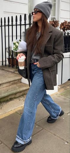 Woman in a gray beanie and plaid blazer walking outdoors with coffee and flowers