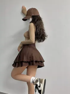 Person in a brown baseball cap and pleated skirt leaning against a wall