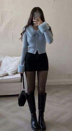 Woman in a blue fur-collared cardigan, black mini skirt, and knee-high boots taking a mirror selfie