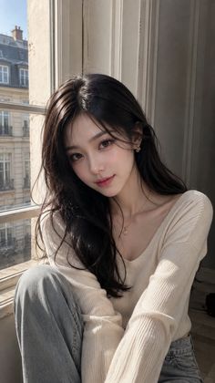 Woman with long dark hair wearing a light sweater and jeans by a window overlooking a city
