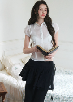 Woman in a white sheer shirt and black skirt holding a book in a bedroom