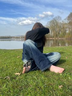 Person with long hair sitting barefoot in grass by a lake