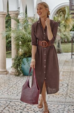 Woman in a brown shirt dress and woven belt, holding a bag outdoors
