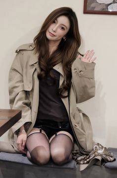 Woman in a trench coat, black shorts, and stockings kneeling indoors