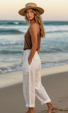 Woman in a straw hat and white pants walking barefoot on a beach at golden hour