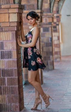 Woman in a black floral dress and high heels leaning against a brick wall
