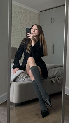 Woman in a black blazer and knee-high socks taking a mirror selfie in a bedroom