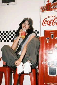 Woman in a baseball cap and cargo pants drinking from a cup next to a Coca-Cola machine