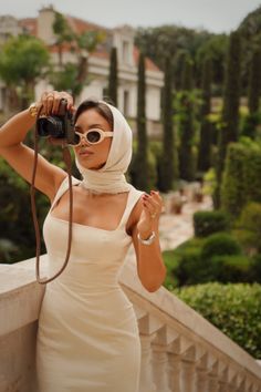 Woman in a cream dress and head scarf holding a camera outdoors