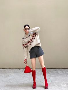 Woman in a fair isle sweater, gray shorts, and red accessories posing outdoors