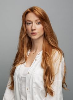 Woman with long red hair and freckles wearing a white shirt against a gray background