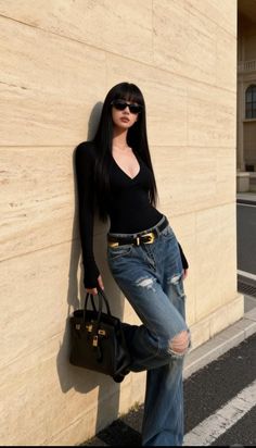 Woman with long black hair and sunglasses leaning against a wall outdoors