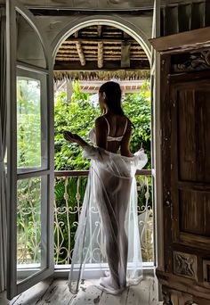 Woman in a white bra and sheer robe standing on a balcony with lush greenery