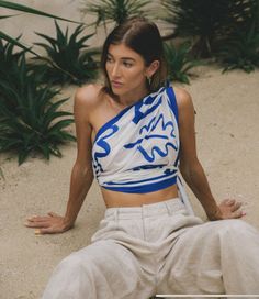 Woman in a one-shoulder top and wide-leg pants sitting on sand with plants