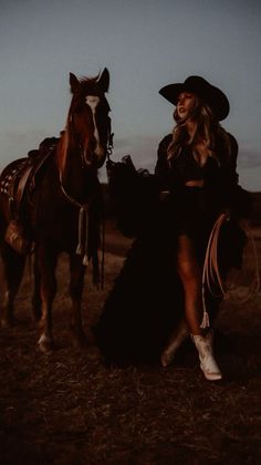 Woman in a cowgirl hat and boots standing next to a horse at night