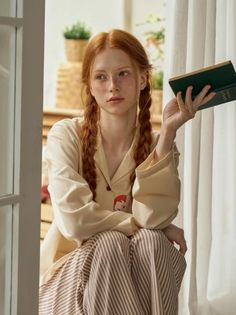 Woman with red braided hair and freckles wearing pajamas, holding a book indoors