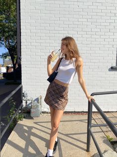 Young woman in a plaid skirt and white tank top drinking a beverage outdoors