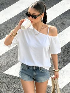 Woman in denim shorts and a white t-shirt drinking from a cup on a street