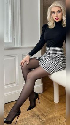 Woman with blonde hair in a black turtleneck and plaid skirt sitting indoors