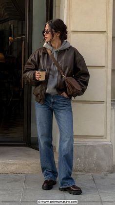 Woman in a brown leather jacket and blue jeans holding a coffee cup outdoors