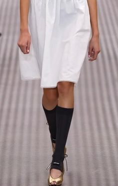 Model in a white dress, black socks, and gold peep-toe heels on a runway