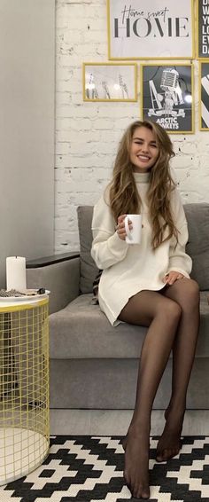 Woman with long hair in a white sweater dress and black tights sitting on a couch