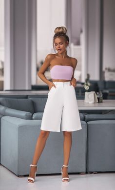 Woman with blonde hair in a high bun wearing a lavender tube top and white shorts indoors