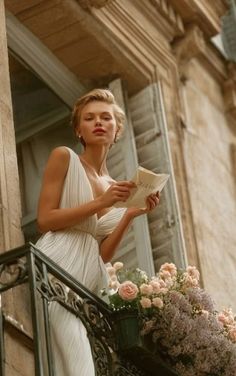 Woman in a white dress on a balcony with flowers, holding a letter