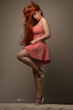 Woman with long red hair in a red gingham dress and high heels posing in a studio