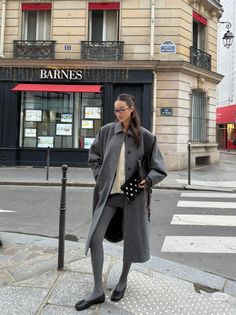 Woman in a long grey coat and glasses standing on a city street