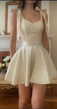 Woman in a cream mini dress with bow straps and a pearl necklace indoors