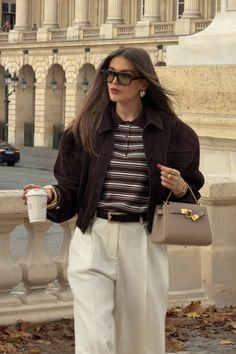Woman in a brown jacket and white pants holding a coffee cup and handbag outdoors