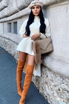 Woman in a white sweater dress and over the knee boots leaning against a wall
