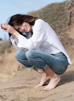 Woman in a white shirt and jeans crouching barefoot on sandy ground