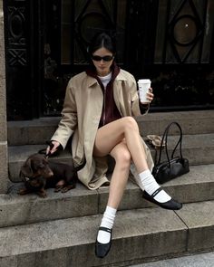 Woman in a trench coat and sunglasses sitting on steps with a dachshund