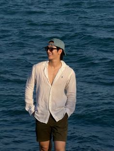 Man in a white shirt, shorts, and sunglasses smiling by the ocean