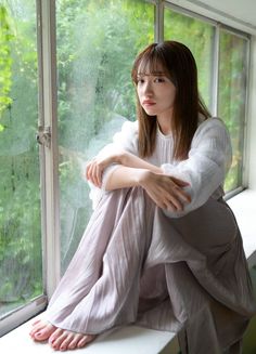 Woman with long hair in a white blouse and pleated skirt sitting by a window