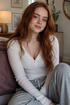 Woman with red hair and freckles wearing a white top and grey sweatpants indoors