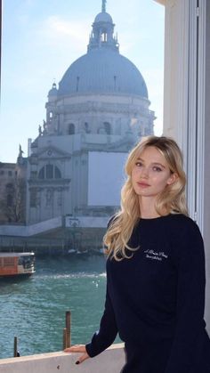 Blonde woman in a navy sweater by a window overlooking a cityscape and water