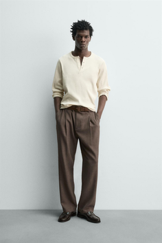 Man in a cream henley shirt and brown pants posing in a studio