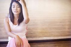 Woman with long hair in a white tank top and pink skirt sitting indoors
